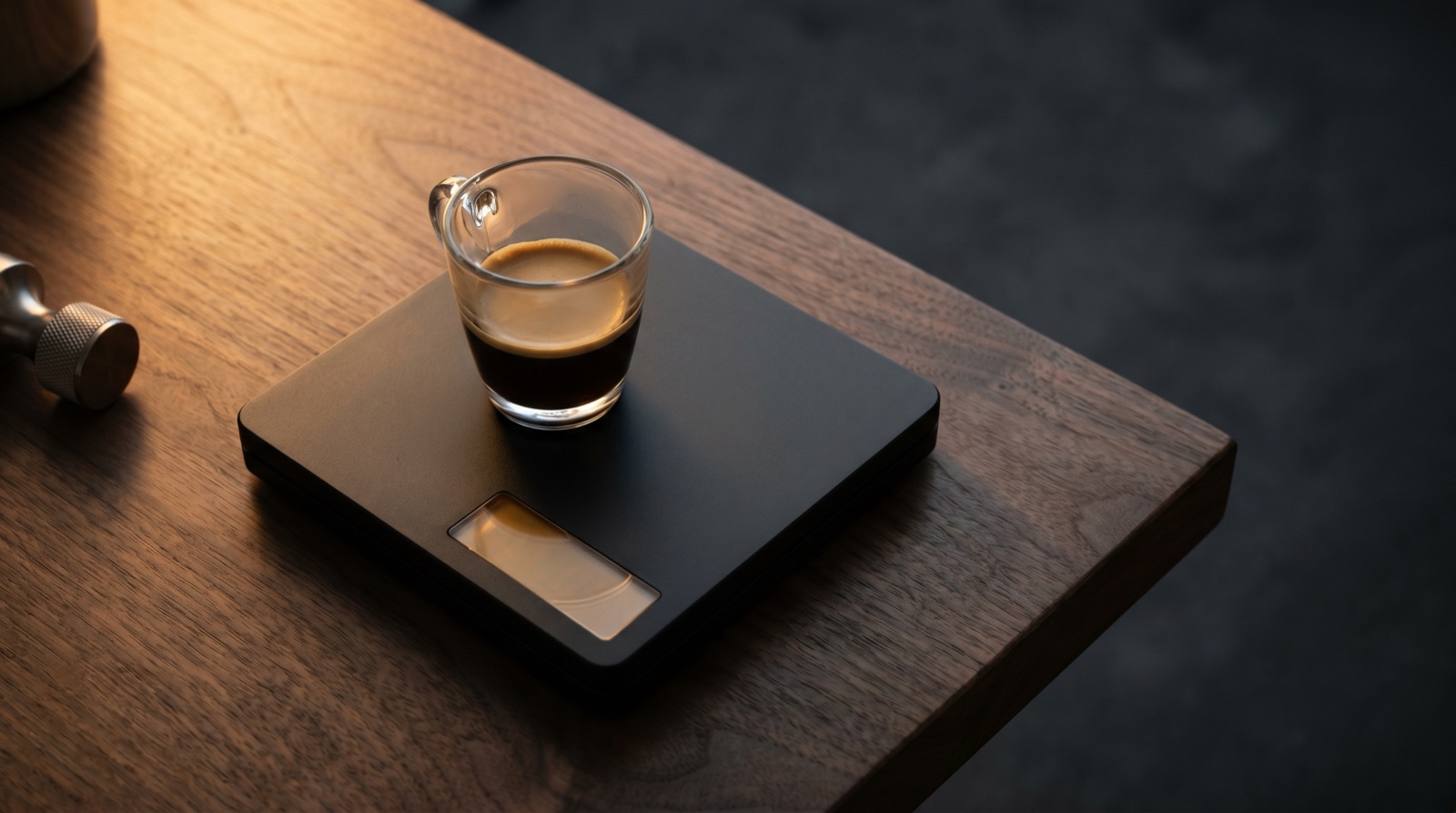 A black digital coffee scale with a glass espresso cup on top, warm side-lighting.