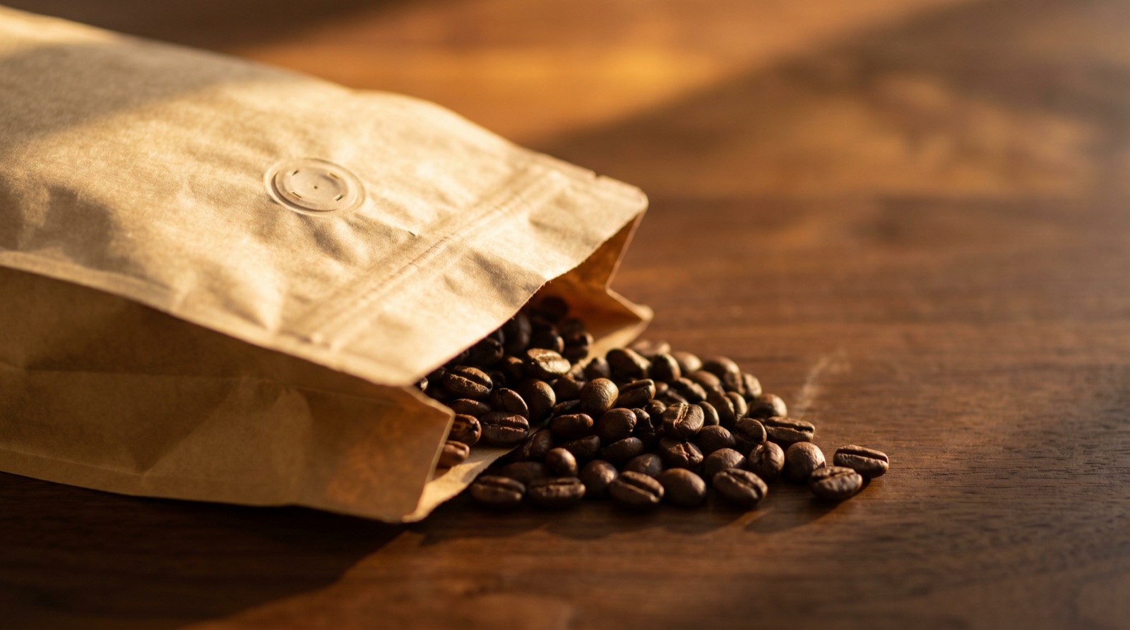 An open kraft-paper coffee bag with whole beans spilling onto dark walnut wood. The bag has a visible one-way degassing valve.