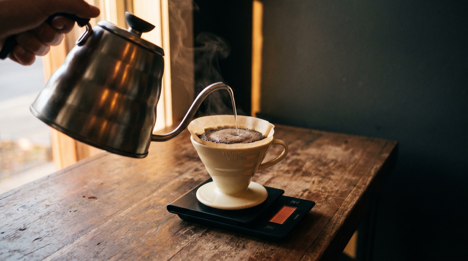 A Hario V60 ceramic dripper mid-pour on a digital scale, gooseneck kettle pouring water, steam rising, warm wood kitchen surface with moody side-lighting.