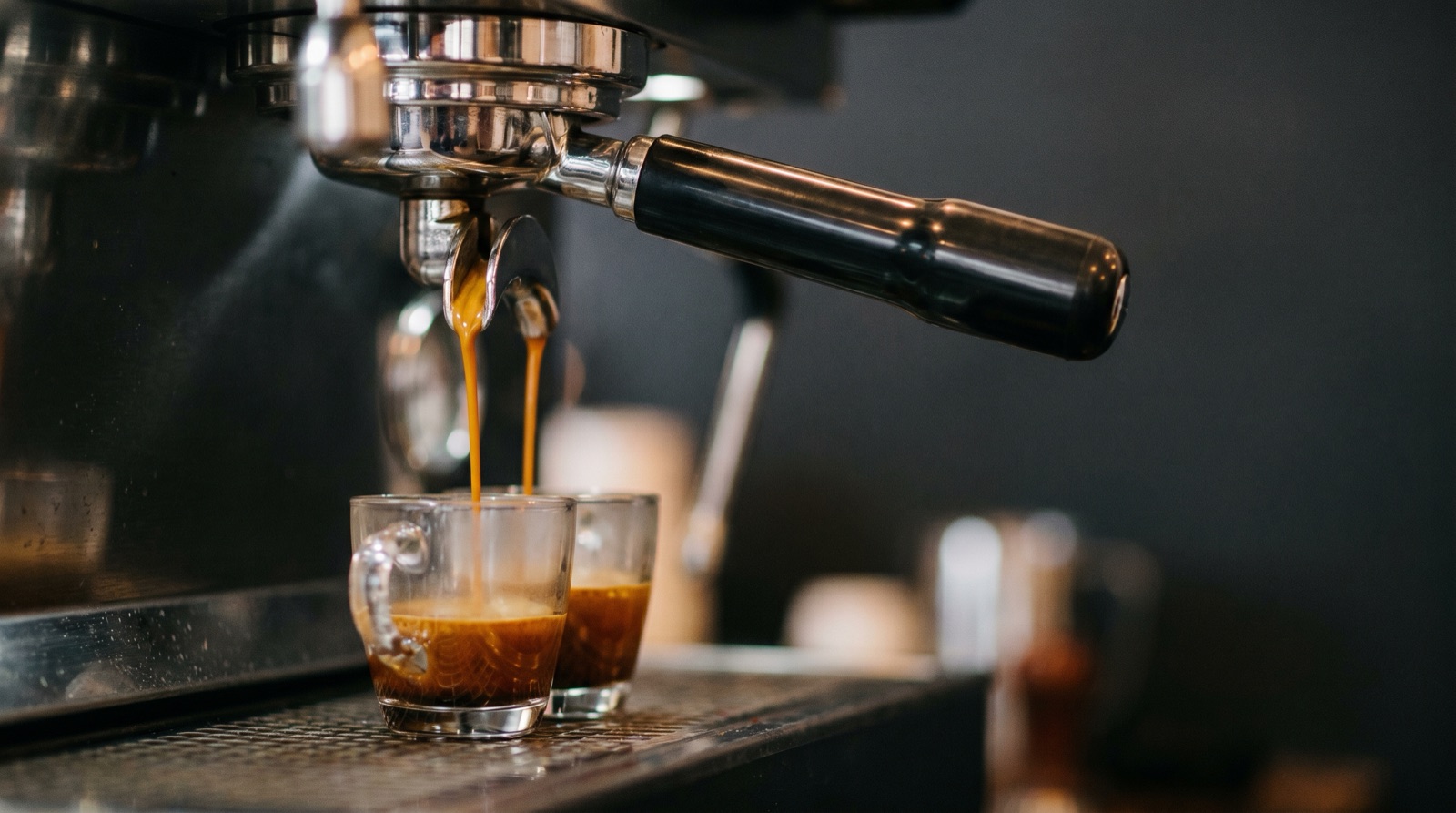 Espresso pulling from a double-spout portafilter into two glass demitasse cups, dark amber crema forming.